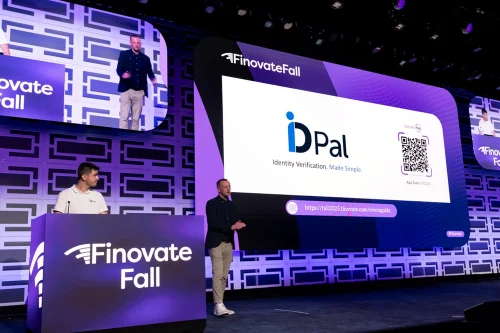 Two presenters from ID-Pal deliver a live demonstration at FinovateFall. One stands behind a podium displaying the Finovate Fall logo, while the other speaks on stage beside a large screen showing the ID-Pal logo, tagline “Identity Verification. Made Simple.”, a QR code, and event details. The backdrop is illuminated in purple and blue tones with a geometric pattern, and overhead screens display the presenters.