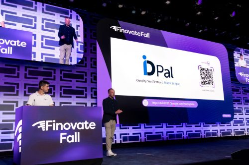 Two presenters from ID-Pal deliver a live demonstration at FinovateFall. One stands behind a podium displaying the Finovate Fall logo, while the other speaks on stage beside a large screen showing the ID-Pal logo, tagline “Identity Verification. Made Simple.”, a QR code, and event details. The backdrop is illuminated in purple and blue tones with a geometric pattern, and overhead screens display the presenters.