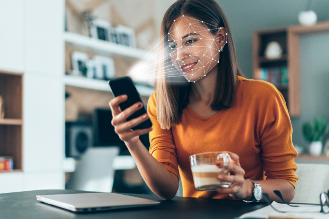 Smiling woman in an orange top sitting at a desk, holding a smartphone in one hand and a glass of milk in the other, with a laptop in front of her.